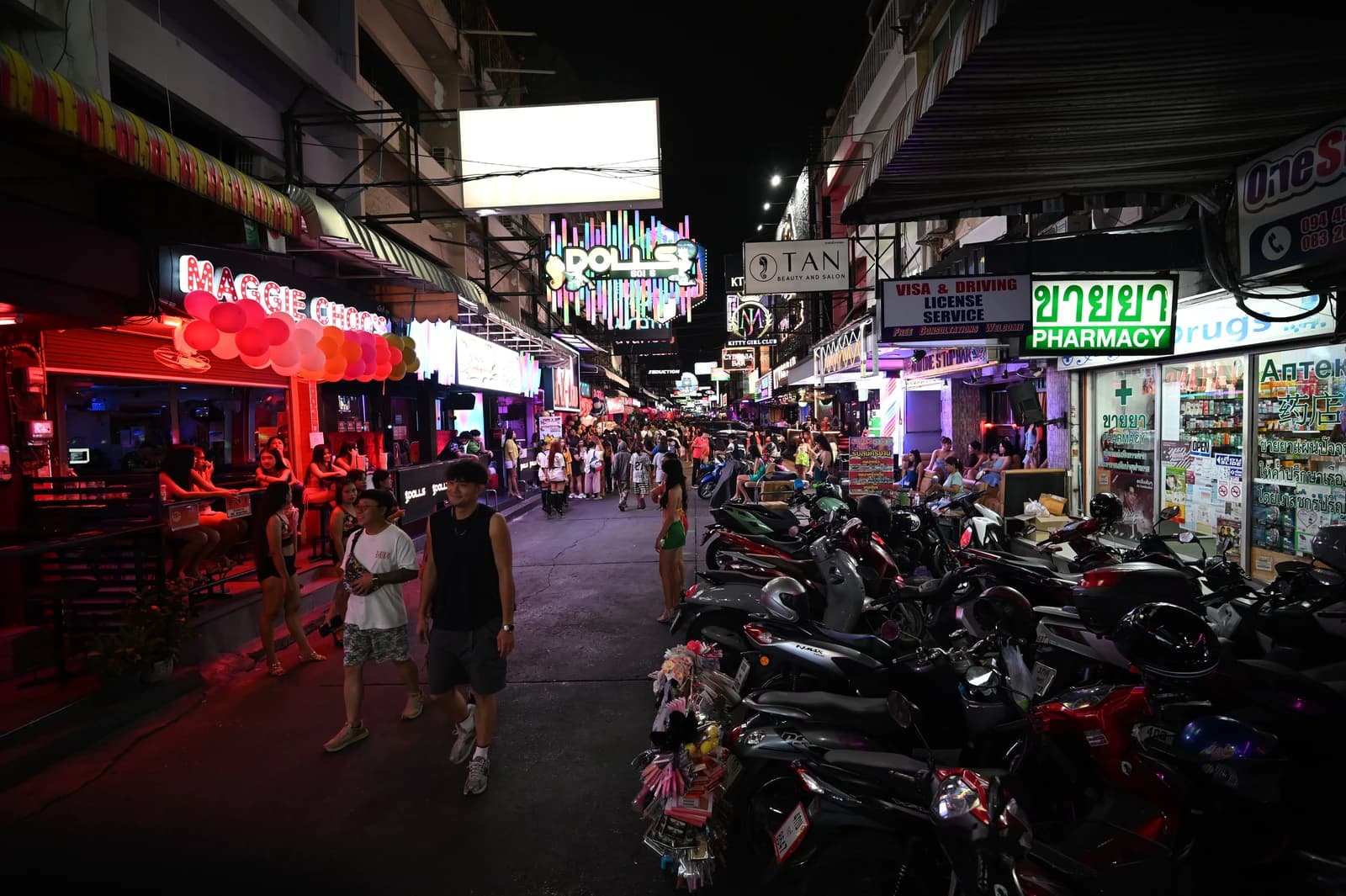 People walking on a busy, neon-lit street with parked scooters in Thailand at night.