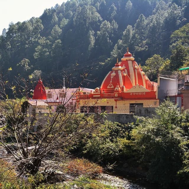 Kainchi Dham Neem Karoli Temple, a red and yellow structure, with trees and a stream.
