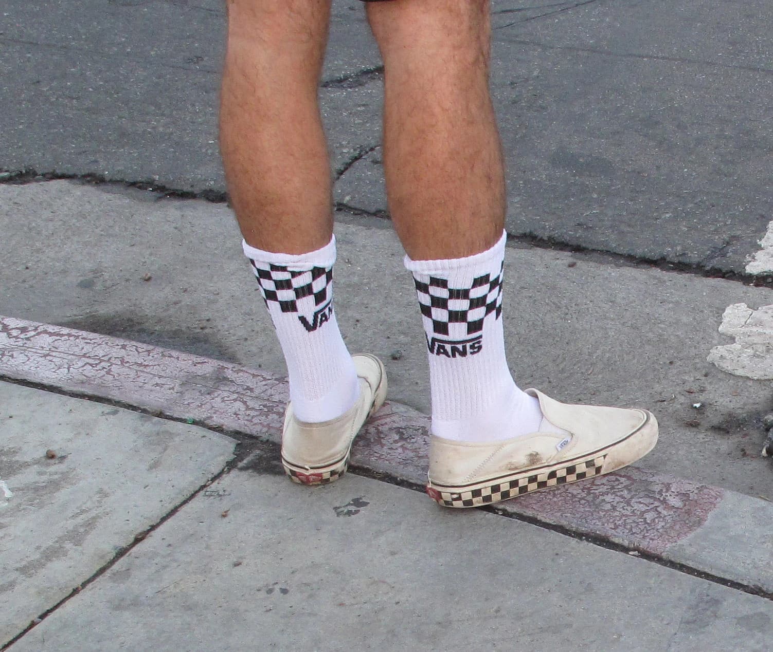 A person stands on a sidewalk wearing white Vans slip-on sneakers and Vans socks.