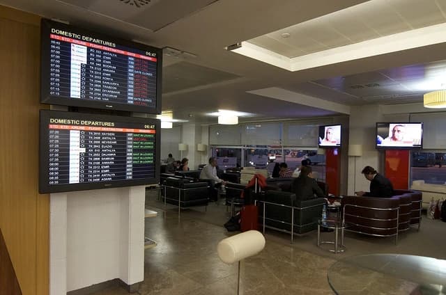Travelers sitting in comfortable chairs in an airport lounge with flight information screens on a wall.