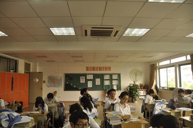Students in a classroom, seated at individual desks, reading books and writing.