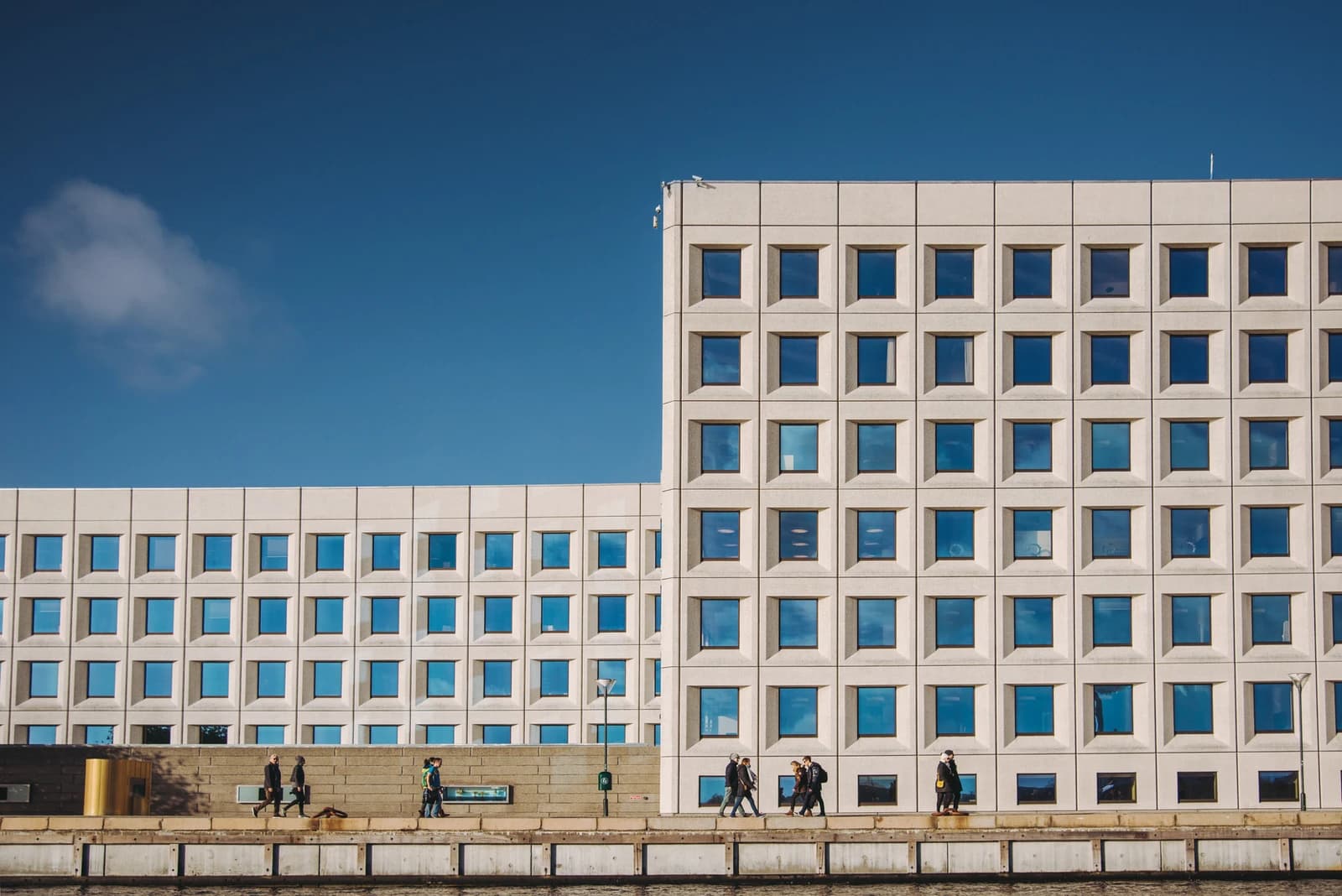 People walking on a path alongside a river with modern office buildings.
