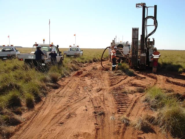 Drill rig on red earth with three workers and several utility vehicles in a flat, grassy landscape.