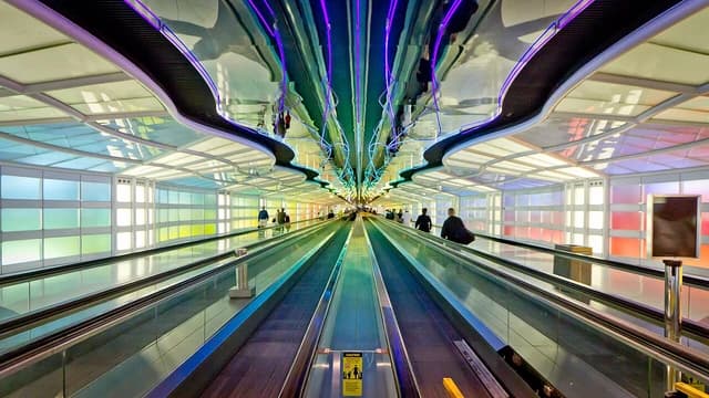 Travelers on moving walkways in a long, brightly illuminated airport concourse.