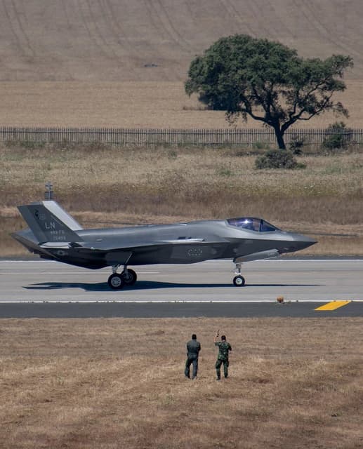 An F-35 fighter jet taxing on a runway with two people observing from the grass.