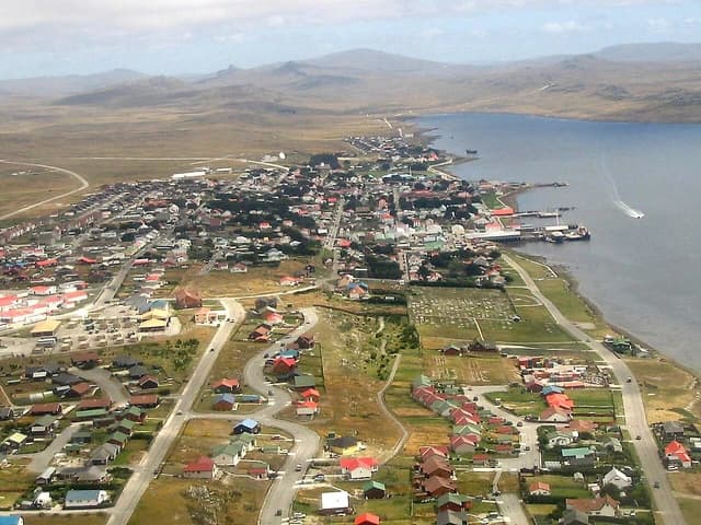 Aerial view of Port Stanley settlement, harbor, and surrounding landscape under cloudy sky.