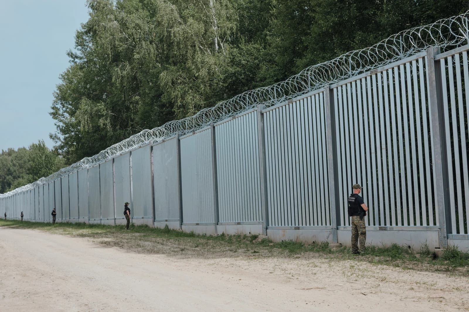 Uniformed personnel standing alongside a tall metal barrier topped with barbed wire.