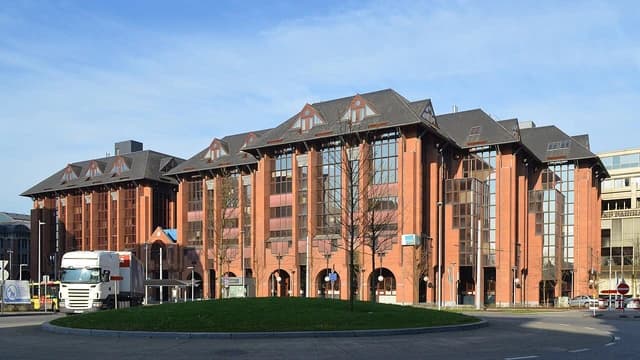Brick building with large windows, surrounded by a road and grassy area.