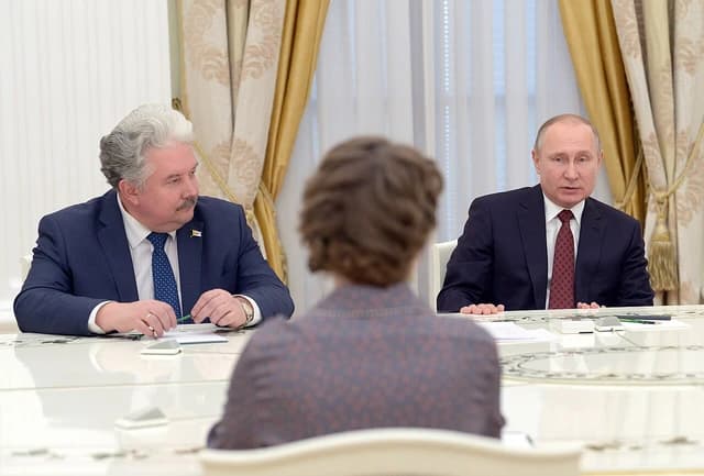 Vladimir Putin seated at a long table opposite another man and a third person with their back to the camera.