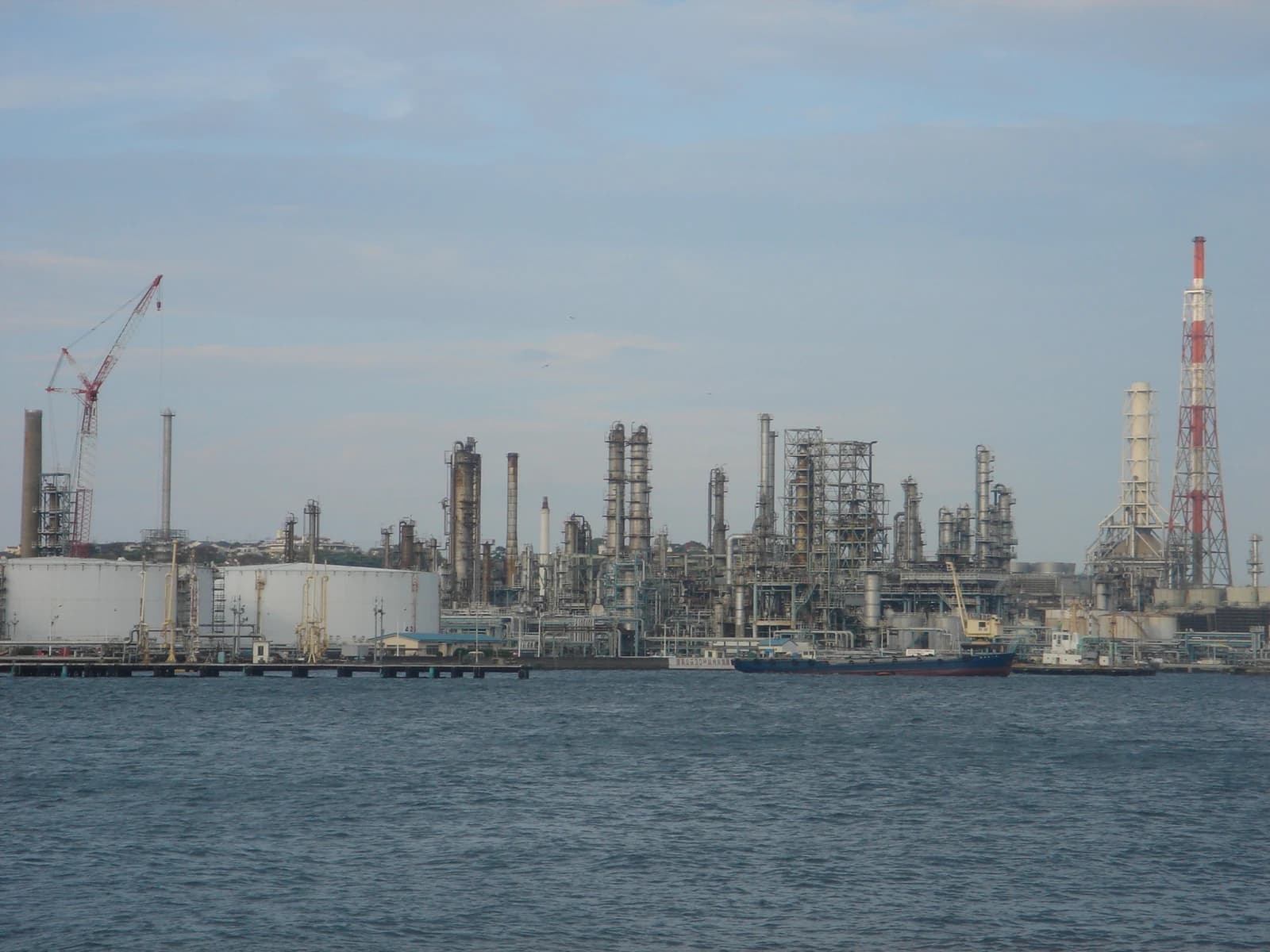 An oil refinery with industrial structures, storage tanks, and a large crane by the water.
