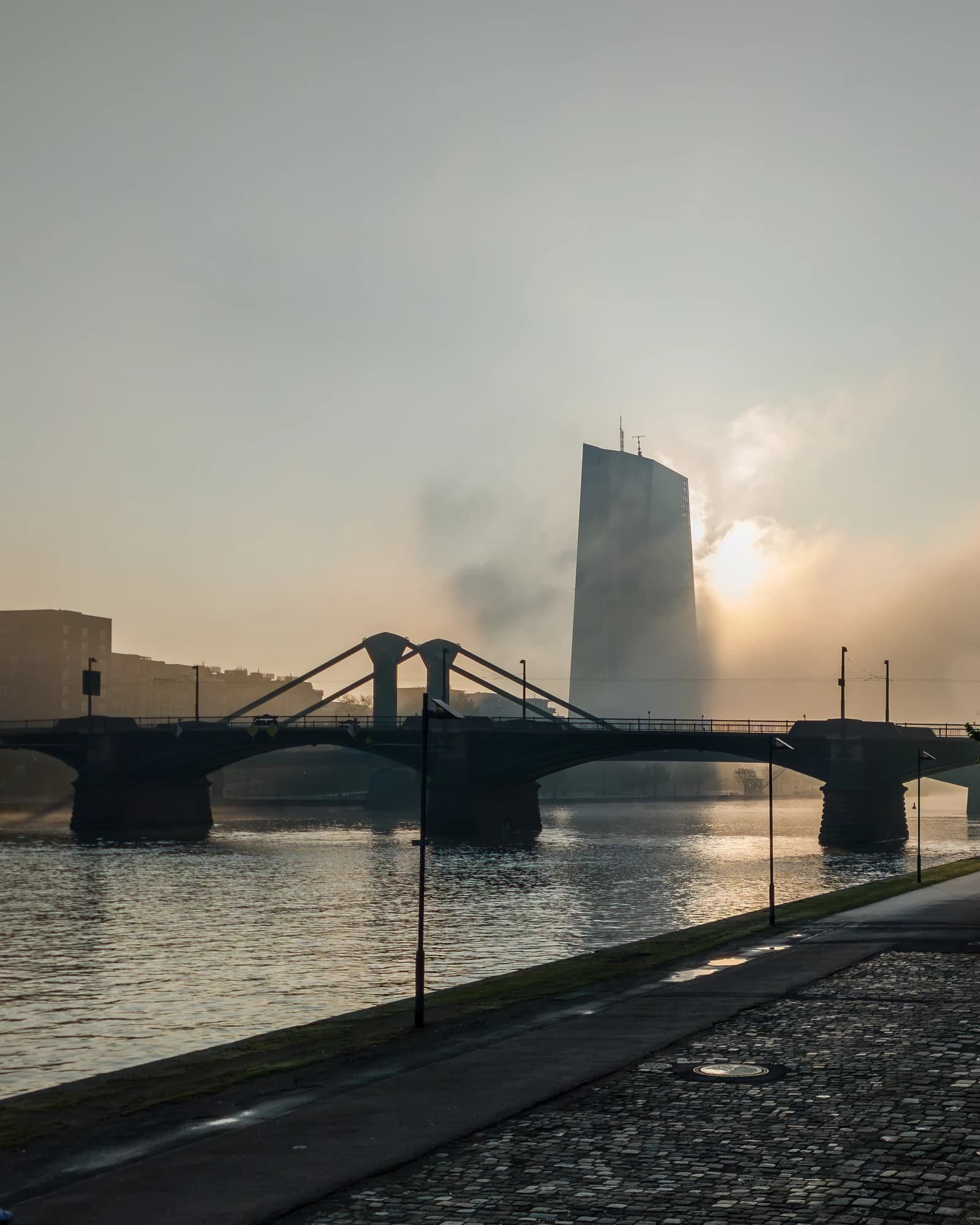 European Central Bank headquarters building partially obscured by mist at sunrise over a river.