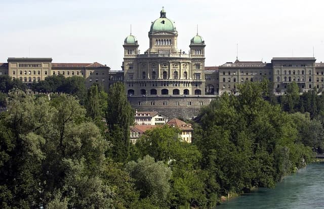 The Swiss Federal Palace, a large domed building, stands above green trees and a river.