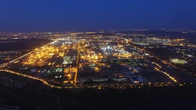 Aerial night view of an illuminated oil refinery with city lights in the background.
