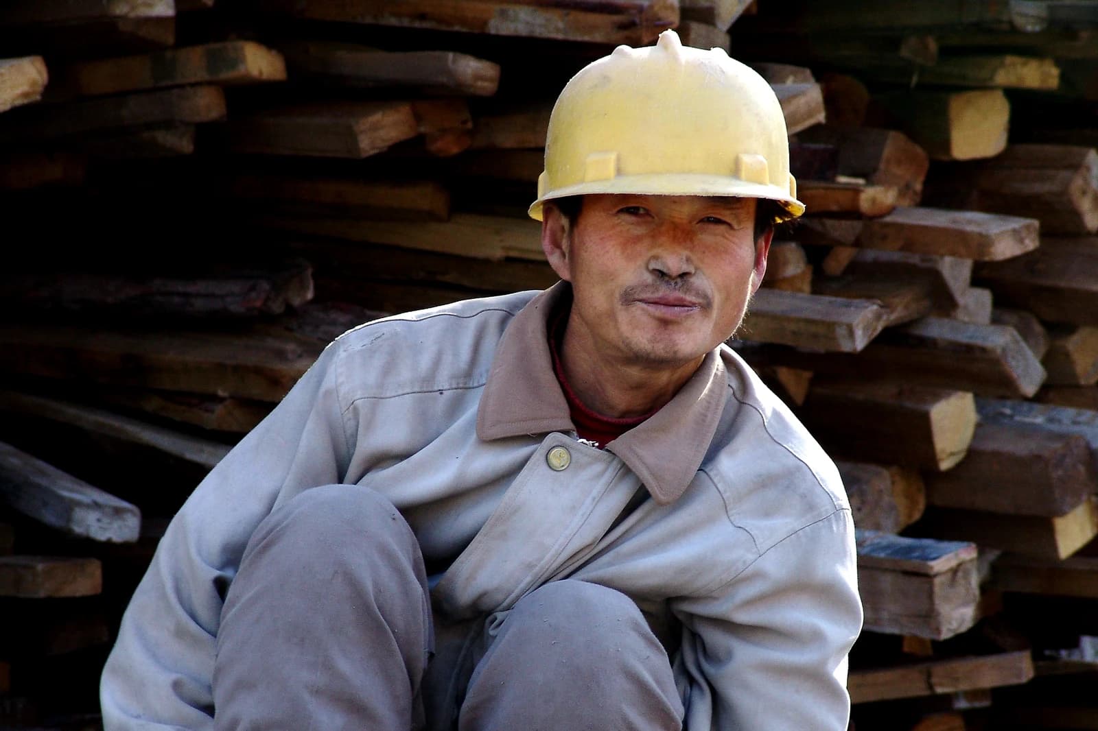 Construction worker wearing yellow hard hat, sitting by stacked wooden planks.