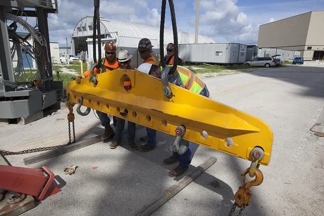 Construction workers position a large yellow lifting beam on an outdoor work site.
