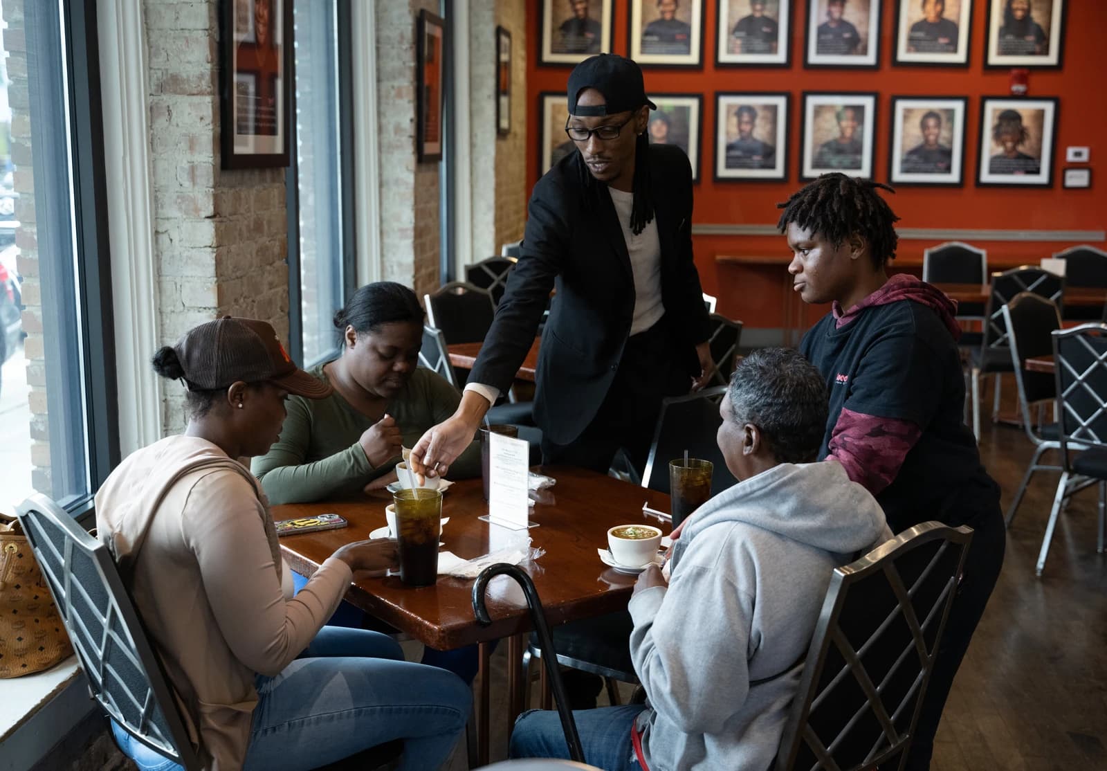 Server in black cap and jacket, serving patrons at a cafe table.