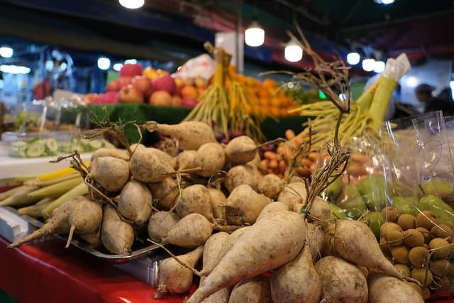 Fresh root vegetables piled on a stall with blurred fruits and produce in the background.