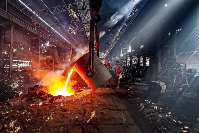 Molten metal flows from a large crucible into a casting bed inside a factory.