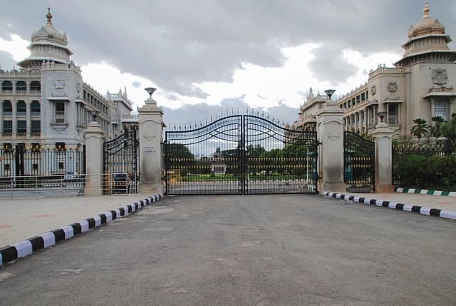 Vidhana Soudha, a large white building with an ornate gate and domed towers, under a cloudy sky.