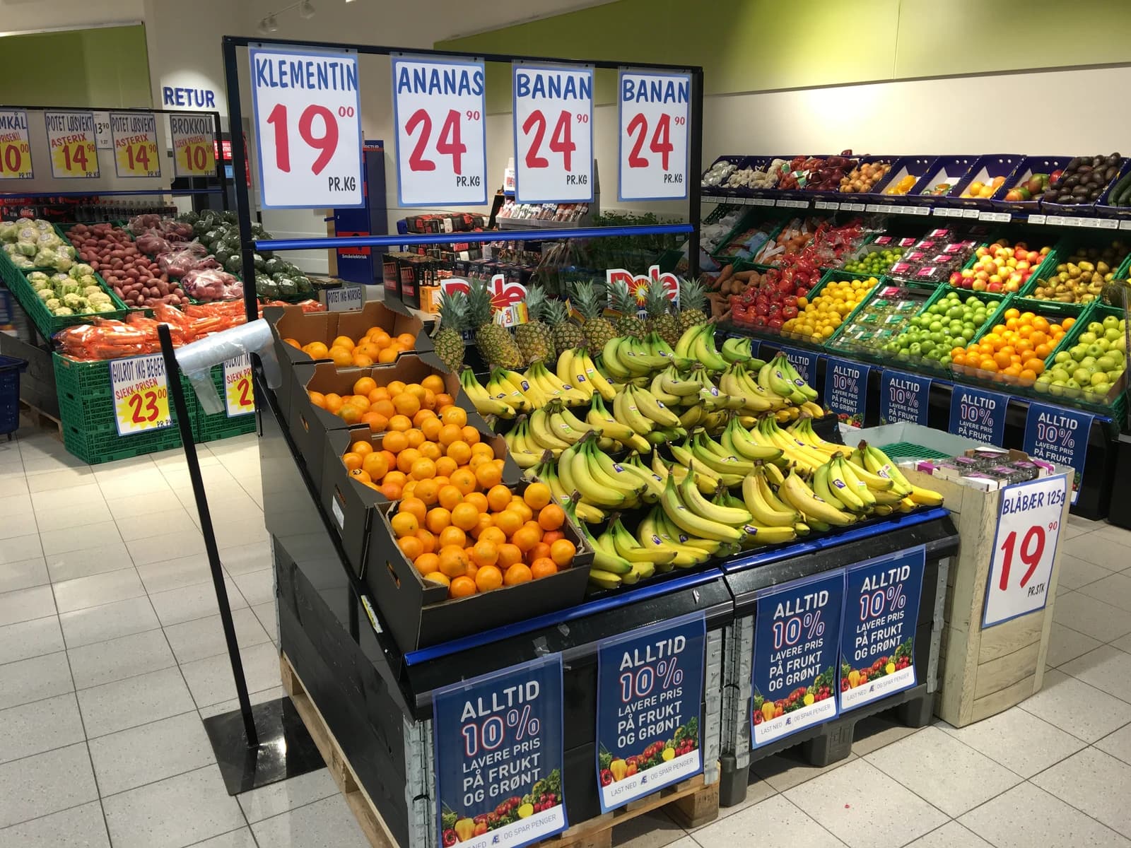 Interior view of a supermarket produce section with displays of oranges, bananas, and other fruits and vegetables.