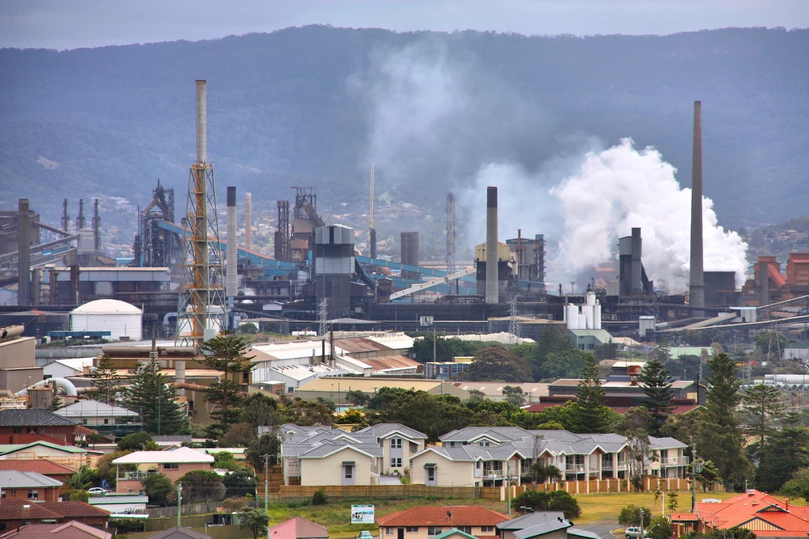 An industrial steelworks facility with multiple chimneys emitting smoke, overlooking a residential area.