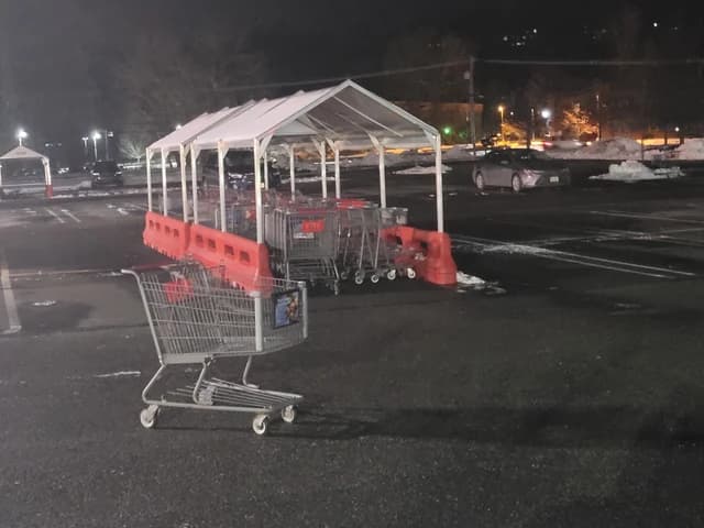 An empty shopping cart stands in a dark, snowy parking lot next to a covered cart return.