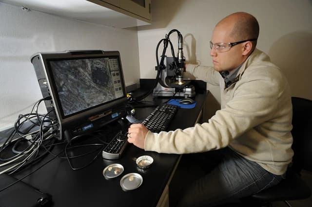 A man wearing safety glasses operates a microscope connected to a computer monitor.