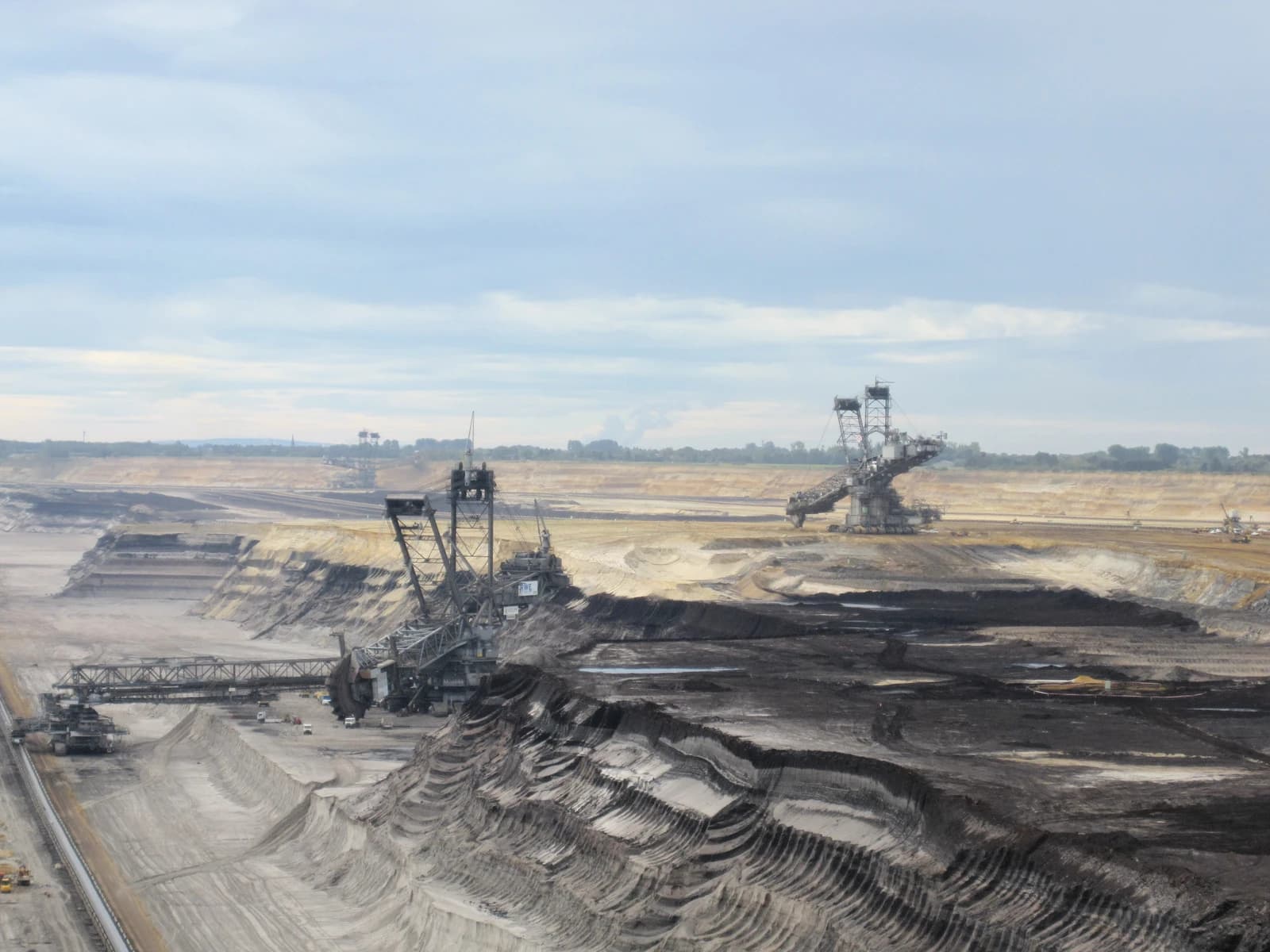 Large industrial excavators operating in a vast open-pit brown coal mine under a cloudy sky.