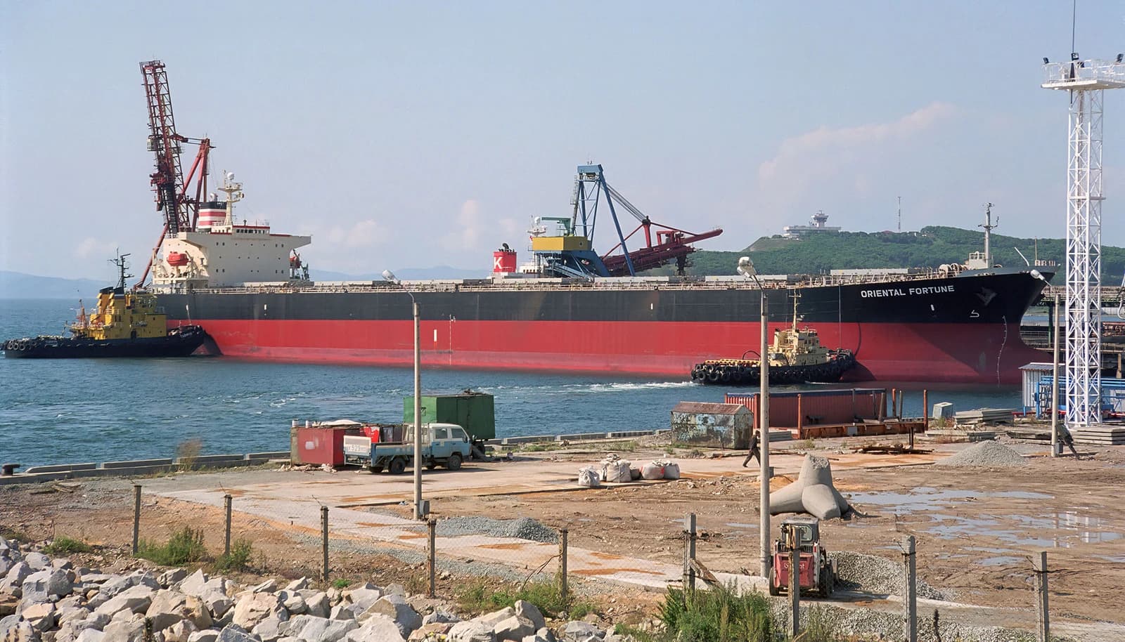 A large bulk carrier ship named 'Oriental Fortune' docked at Vostochny Port, with tugboats alongside.