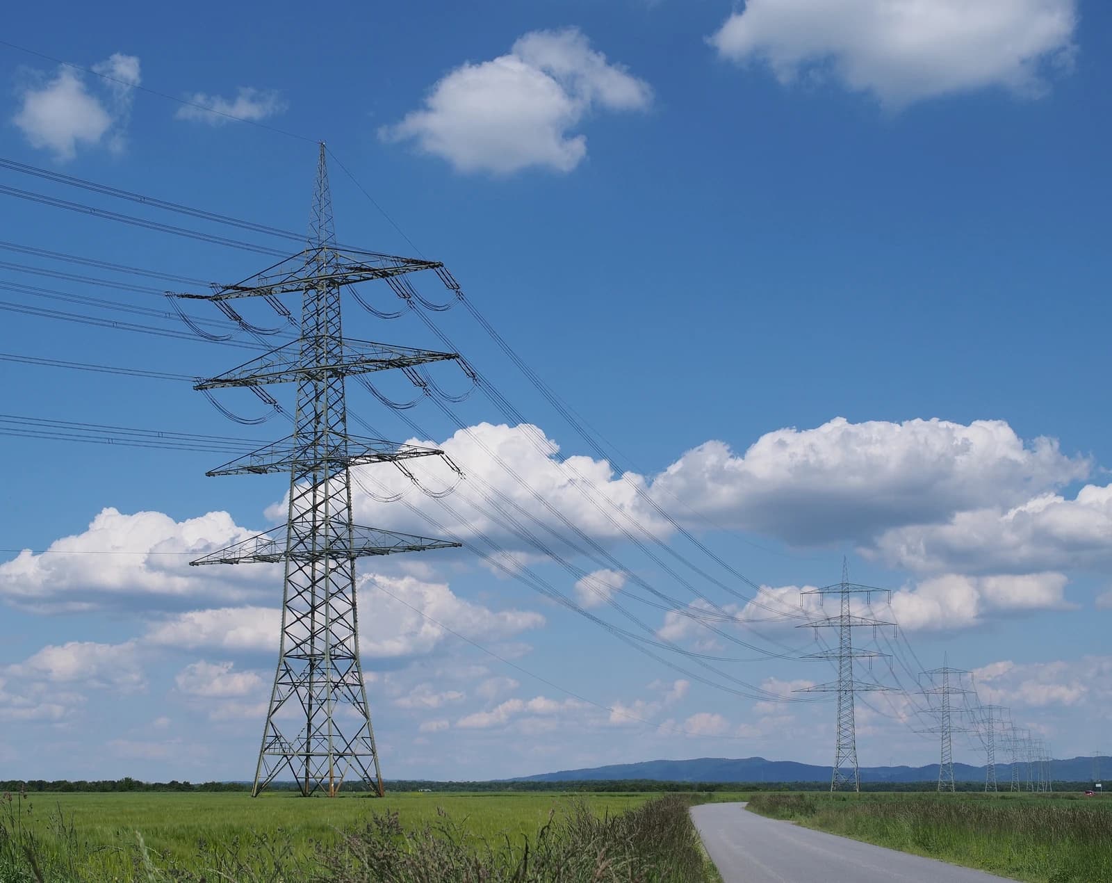 Electricity pylons stretching across a landscape with a winding road and a blue sky with clouds.