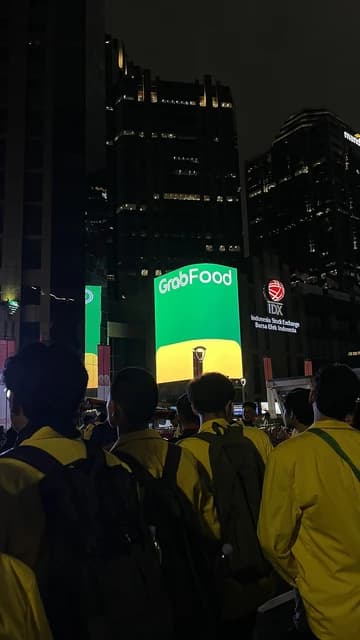 People in yellow jackets and backpacks look towards large illuminated GrabFood and IDX signs at night.
