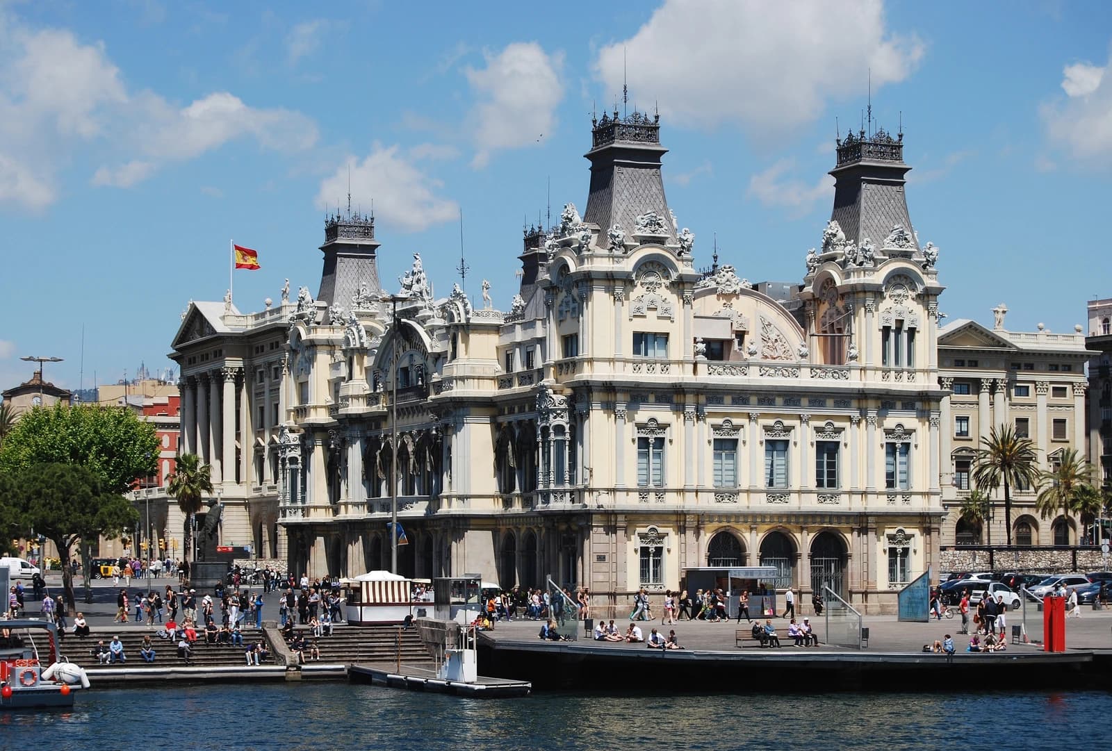 The ornate Port Authority building in Barcelona with a Spanish flag on top, viewed from the water.