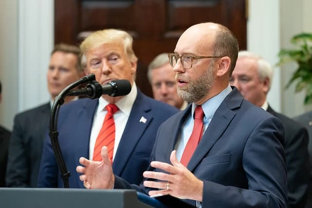 President Trump watching another man speak at a podium with a microphone.