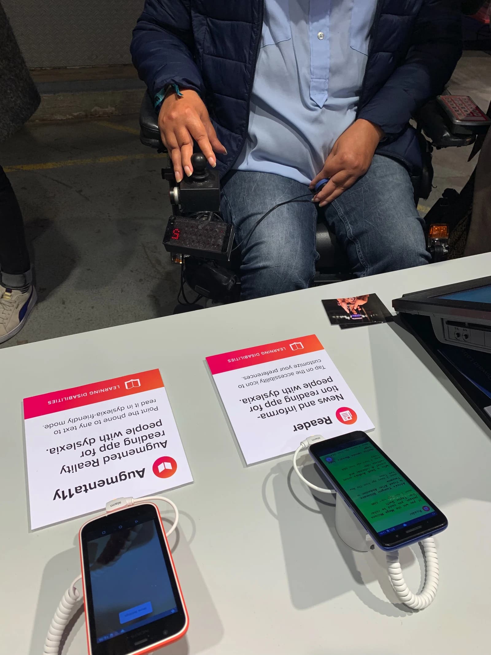 Person in a wheelchair with hands near joystick, next to a table displaying phones with accessibility apps.
