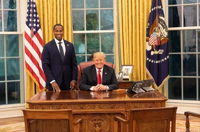 Donald Trump seated at the Resolute Desk with John Edward James standing beside him.