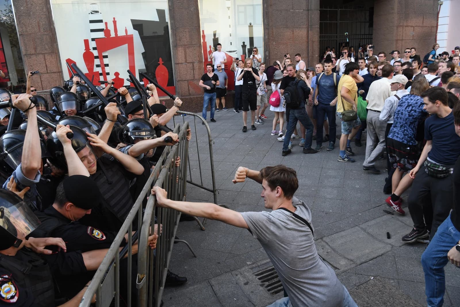 Police officers in riot gear hold batons and metal barriers against protesters in Moscow.