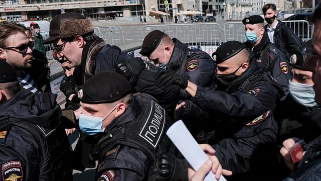 Russian police officers in black uniforms detaining a man in a brown hat and glasses.