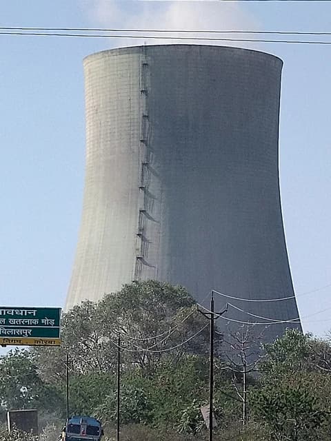 A large concrete cooling tower rises above trees, with power lines and a road sign visible.