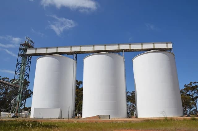 Three large white wheat silos with a loading structure under a bright blue sky.