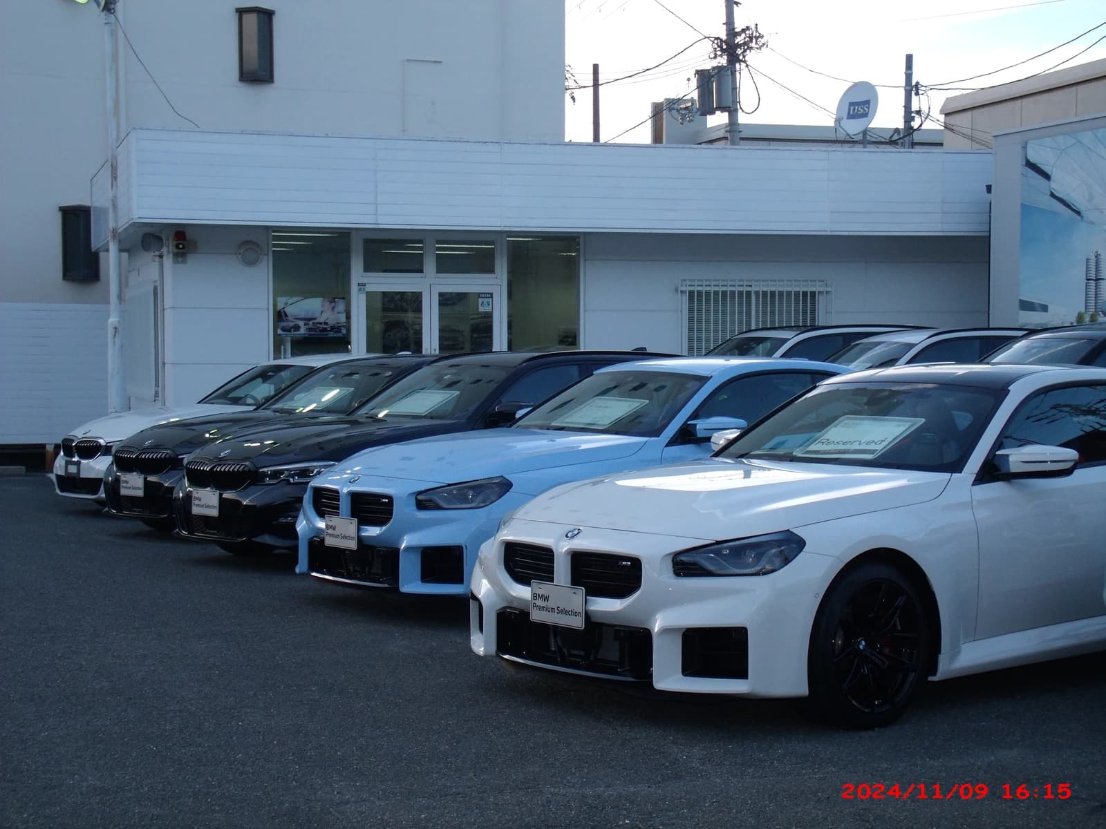 Multiple BMW cars, including white, blue, and black models, lined up outside a dealership building.