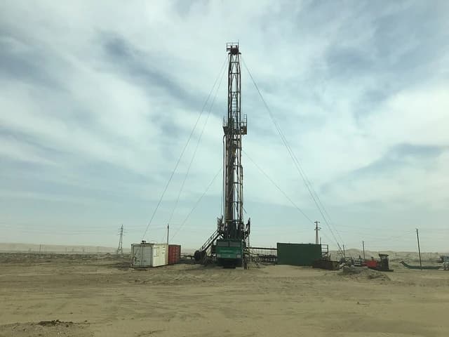 An oil drilling rig stands in a barren desert landscape under a cloudy sky.