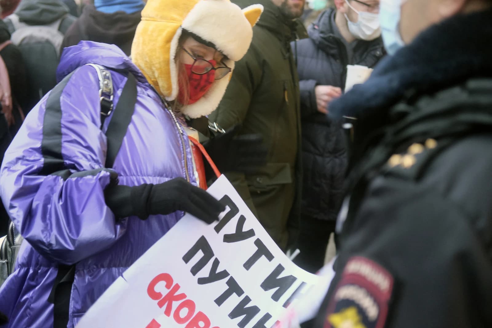 Protester wearing a yellow hat and purple coat holds a sign in Russian, near a police officer.