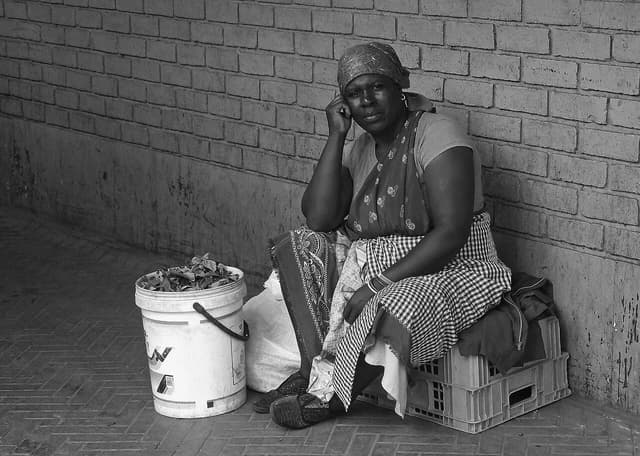 Woman selling fresh produce sits on crates next to a white bucket of leaves.