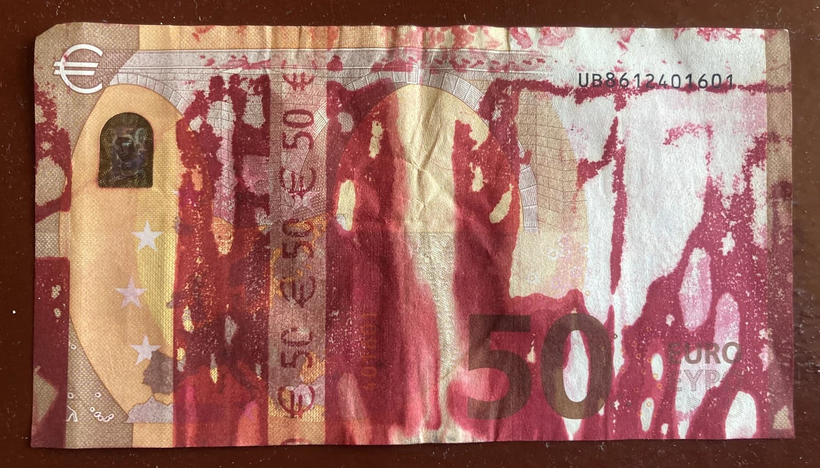 A fifty euro banknote, partially stained with red, resting on a dark brown surface.