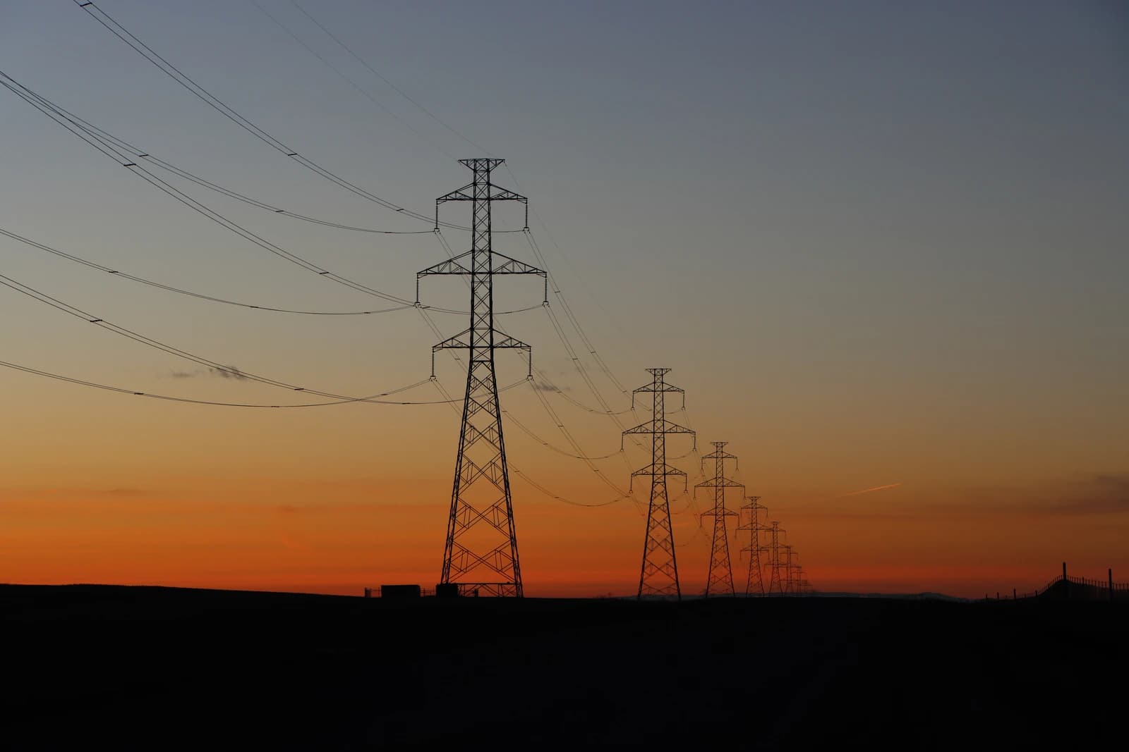 Electricity transmission lines and towers silhouetted against an orange and blue sunset sky.