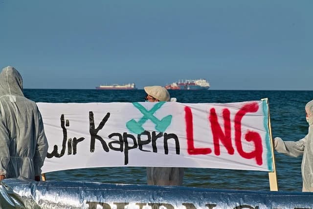 Protesters holding a banner with German text against LNG on a beach in front of LNG tankers.