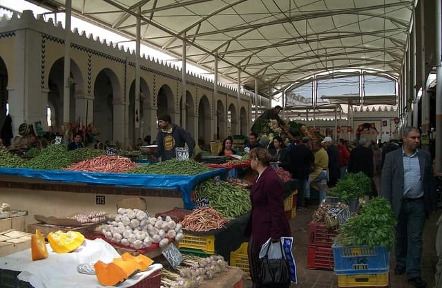 People walking through a bustling market with stalls piled high with fresh vegetables.