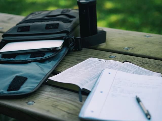 Laptop in a bag, open textbook, and spiral notebook with a pen on a table.