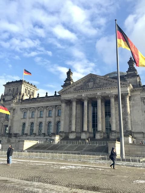 Exterior of the Reichstag building in Berlin with German flags and people walking outside.