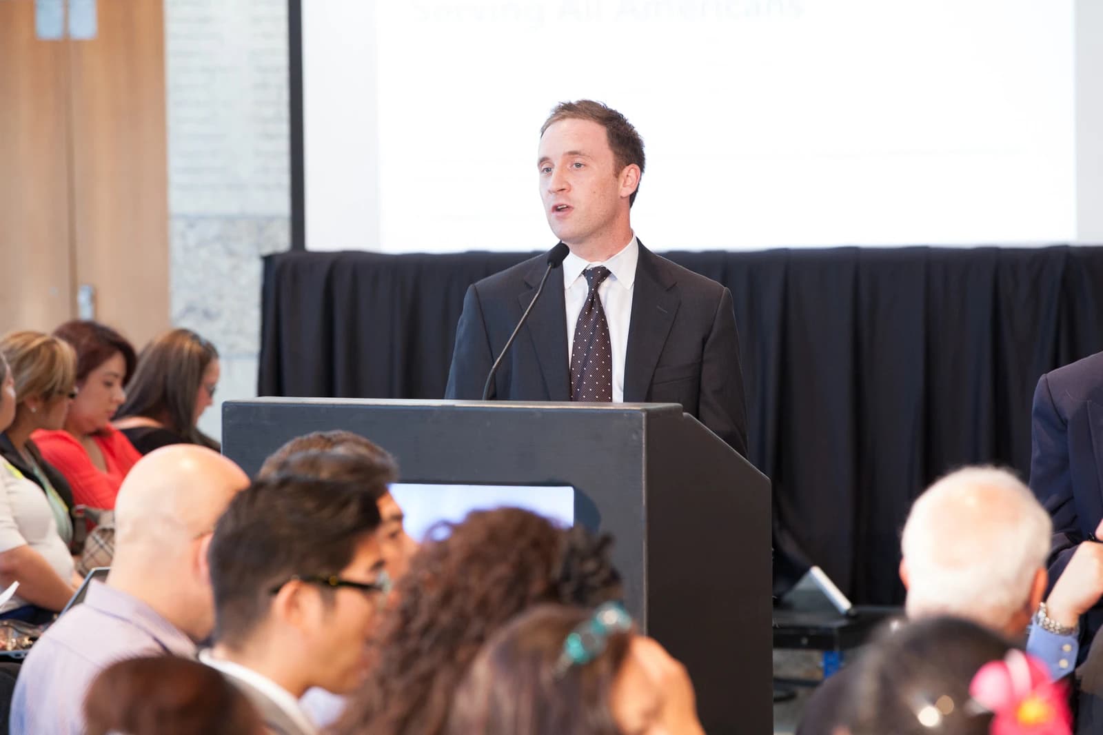 Man in a suit speaks at a podium to an audience in a conference setting.